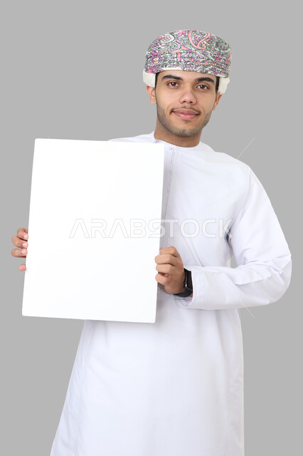 Portrait of an Arab Omani Gulf man in traditional dress, making different facial gestures, holding a white board in his hand, an empty billboard, a gray background.