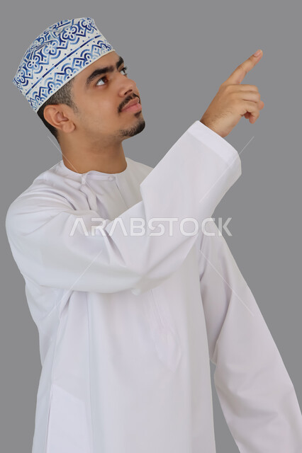 بورتريه لرجل عربي خليجي عماني يشير باصبع السبابة الى اعلى، الاشارة الى شيء ما، حركة اصبع واشارات، خلفية رمادية