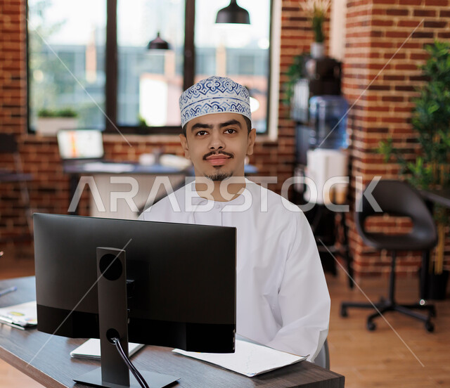رجل عربي خليجي عماني يجلس امام شاشة الحاسوب، انجاز مهام العمل، وظائف مكتبية، اعمال مكتبية، مقر العمل، بيئة عمل، أعمال حره، مكتب عمل