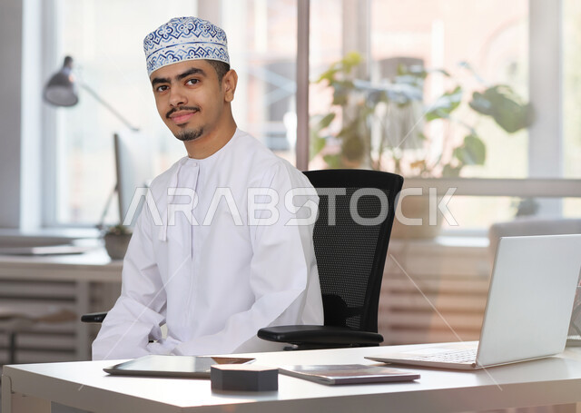 رجل عربي خليجي عماني يجلس في مكتب العمل، استخدام جهاز الحاسوب المحمول، انجاز مهام العمل، تصفح الانترنت، احتراف وظائف الحاسوب وتكنولوجيا المعلومات، اعمال مكتبية، مقر العمل، استخدام وسائل تقنية حديثة