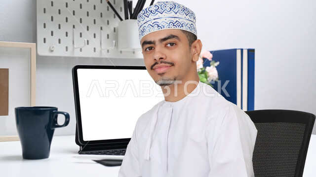 An Arab Gulf Omani man sitting in the work office, using a laptop, completing work tasks, surfing the Internet, professionalizing computer and information technology jobs, office work, workplace, using modern technical means