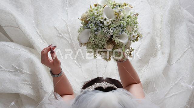 صورة من الأعلى لعروس عربية خليجية سعودية تقوم بجلسة تصوير ، مسكة عروس يوم زفافها ، بدلة زفاف بيضاء ، باقة ورود ، ليلة العمر ، مناسبات وأفراح .
