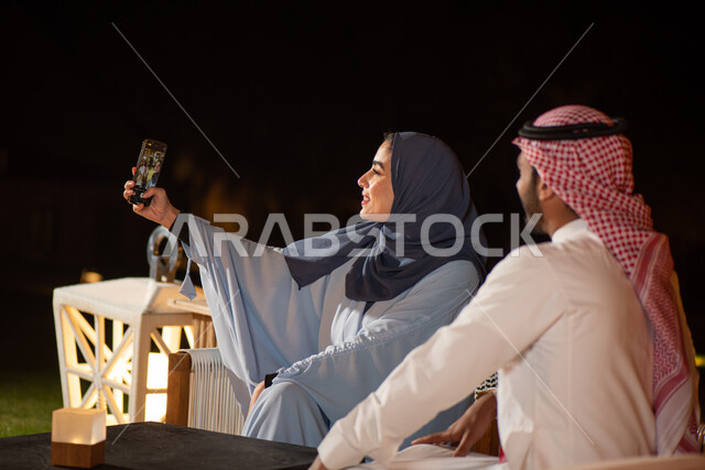 زوجان عربيان خليجيان سعوديان يستخدامان الهاتف المحمول، تقنيات حديثة، قضاء اوقات ممتعة، التقاط صورة تذكارية، صور سيلفي، الكاميرا الامامية، وناسة وتعارف، الجلوس في الهواء الطلق