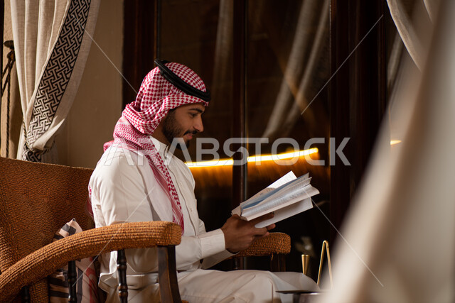 رجل عربي خليجي سعودي يحمل بيده كتاب ويجلس امام النافذة، قضاء الوقت بالقراءة، التعلم والثقافة، القراءة والمعرفة، جلسة سعودية