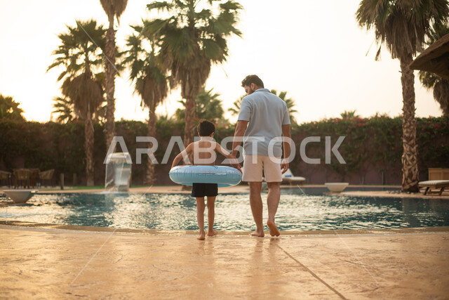 A Saudi Arabian Gulf man spending fun times with his son, walking on the water pond borders, using a swimming buoy, getting ready to swim in the pool, a pleasant summer atmosphere, a recreational tourist trip, a happy summer vacation
