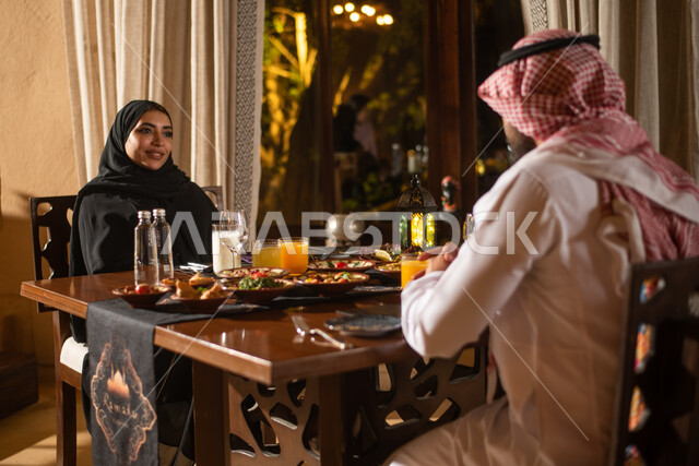 زوجان عربيان خليجيان سعوديان على مائدة الافطار بشهر رمضان، تناول وجبة الافطار، سفرة مليئة باصناف من المأكولات اللذيذة، ايماءات وجه سعيدة، اجواء رمضانية، فانوس رمضان، شهر رمضان الكريم