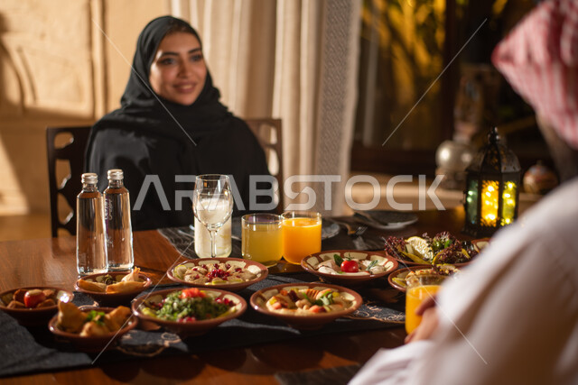 زوجان عربيان خليجيان سعوديان على مائدة الافطار بشهر رمضان، تناول وجبة الافطار، ايماءات وجه سعيدة، سفرة مليئة باصناف من المأكولات اللذيذة، اجواء رمضانية، فانوس رمضان، شهر رمضان الكريم