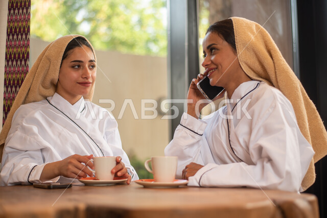 امرأتان عربيتان خليجيتان سعوديتان تستمتعان بشرب المشروبات الساخنة، منتجع صحي نسائي، استخدام الهاتف المحمول، اجراء مكالمة هاتفية، اتصال وتواصل، دردشة ومحادثات، الراحة والاستجمام، الاهتمام والعناية بالمظهر، صالون وسبا نسائي
