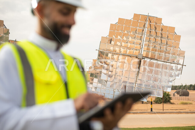 مهندس طاقة عربي خليجي سعودي يحمل بيده جهاز تقني، إرتداء خوذة وسترة الحماية الخاصة بالعمل، التدقيق والإشراف على العمل، رؤية السعودية 2030، الطاقة المتجددة، الطاقة النظيفة، الطاقة الطبيعية، ستلايت، محطات الطاقة الشمسية