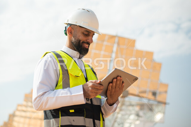 مهندس طاقة عربي خليجي سعودي يحمل بيده جهاز تقني، التدقيق والإشراف على العمل، إرتداء خوذة وسترة الحماية الخاصة بالعمل، رؤية السعودية 2030، الطاقة المتجددة، الطاقة النظيفة، الطاقة الطبيعية، ستلايت، محطات الطاقة الشمسية