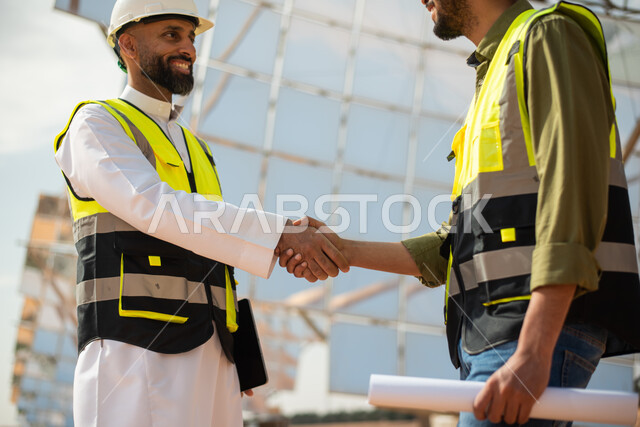 مهندسان طاقة عربيان خليجيان سعوديان يتصافحان، إرتداء خوذة وسترة الحماية الخاصة بالعمل، الاشراف على محطات الطاقة الشمسية في السعودية، مخططات وتصاميم هندسية، حوارات ونقاشات، الطاقة المتجددة، الطاقة النظيفة، الطاقة الطبيعية، ستلايت، محطات الطاقة الشمسية