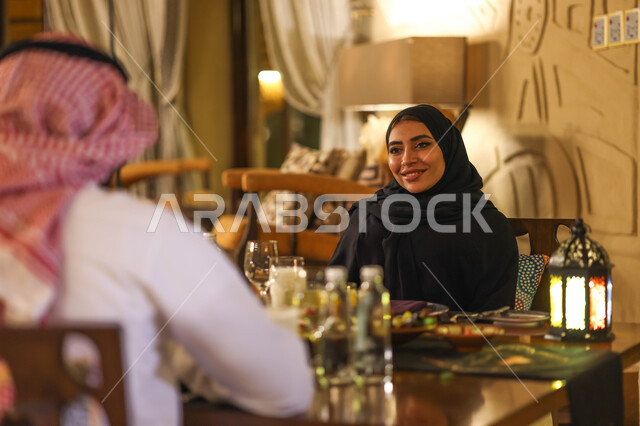 زوجان عربيان خليجيان سعوديان على مائدة الافطار بشهر رمضان، سفرة مليئة باصناف من المأكولات اللذيذة، تناول وجبة الافطار، ايماءات وجه سعدية، اجواء رمضانية، فانوس رمضان، شهر رمضان الكريم