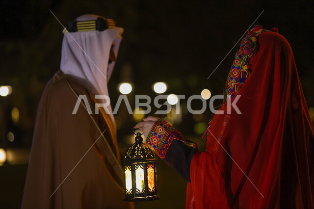 An Arab, Saudi, Gulf couple, dressed in a traditional Saudi folk costume, preparing for the advent of the blessed month of Ramadan, lanterns for the month of Ramadan, lighting and decorations for the month of Ramadan, decorations and lights, Ramadan atmos
