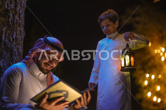 رجل عربي خليجي سعودي يمسك المصحف الشريف وبجانبه ابنه يحمل فانوس رمضان، قراءة وتلاوة القرآن الكريم، حفظ القران الكريم، شهر رمضان المبارك، ايماءات وجه سعيدة، إضاءة وزينة شهر رمضان، أجواء رمضانية، مناسبات دينية