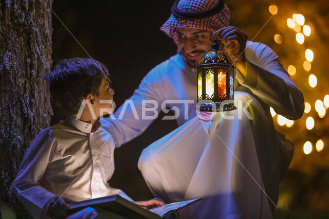 ولد عربي خليجي سعودي يمسك المصحف الشريف وبجانبه والده يحمل فانوس رمضان، قراءة وتلاوة القرآن الكريم، حفظ القران الكريم، شهر رمضان المبارك، ايماءات وجه سعيدة، إضاءة وزينة شهر رمضان، أجواء رمضانية، مناسبات دينية