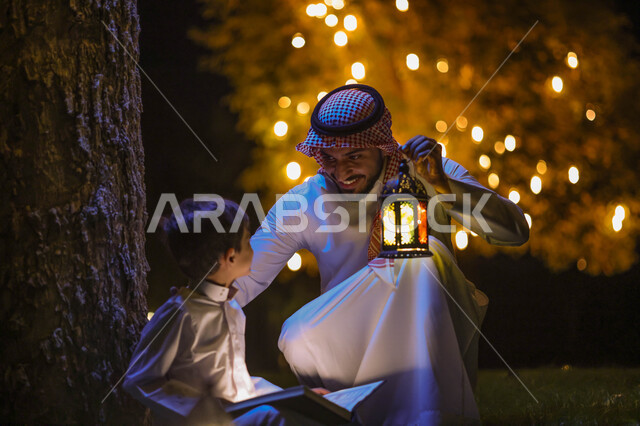 ولد عربي خليجي سعودي يمسك المصحف الشريف وبجانبه والده يحمل فانوس رمضان، قراءة وتلاوة القرآن الكريم، حفظ القران الكريم، شهر رمضان المبارك، ايماءات وجه سعيدة، إضاءة وزينة شهر رمضان، أجواء رمضانية، مناسبات دينية