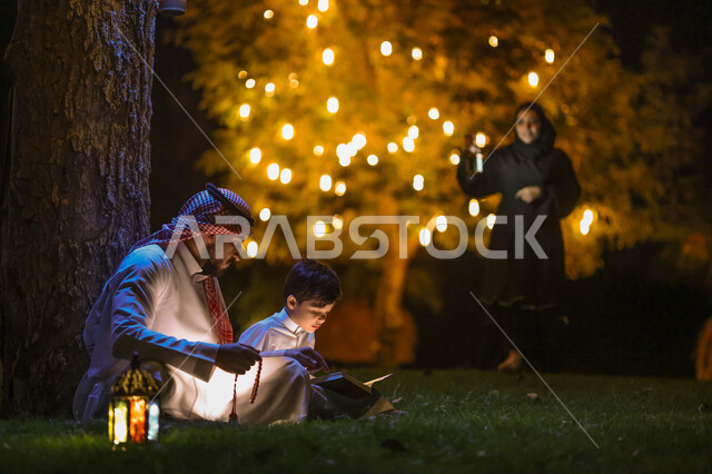 رجل عربي خليجي سعودي يعلم ابنه قراءة وتلاوة القرآن الكريم، تحفيظ القرآن للابناء، تجمع العائلة في حديقة المنزل، شهر رمضان المبارك، فانوس رمضان، إضاءة وزينة شهر رمضان، أجواء رمضانية، مناسبات دينية