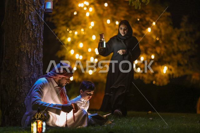 رجل عربي خليجي سعودي يعلم ابنه قراءة وتلاوة القرآن الكريم، تحفيظ القرآن للابناء، تجمع العائلة في حديقة المنزل، شهر رمضان المبارك، فانوس رمضان، إضاءة وزينة شهر رمضان، أجواء رمضانية، مناسبات دينية