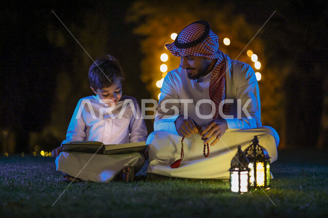 رجل عربي خليجي سعودي يعلم ابنه قراءة وتلاوة القرآن الكريم، تحفيظ القرآن للابناء، شهر رمضان المبارك، فانوس رمضان، إضاءة وزينة شهر رمضان، أجواء رمضانية، مناسبات دينية