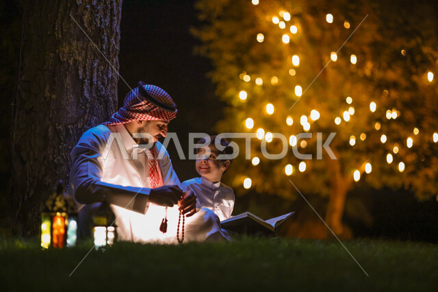 رجل عربي خليجي سعودي يعلم ابنه قراءة وتلاوة القرآن الكريم، تحفيظ القرآن للابناء، شهر رمضان المبارك، فانوس رمضان، إضاءة وزينة شهر رمضان، أجواء رمضانية، مناسبات دينية