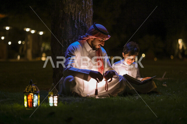 رجل عربي خليجي سعودي يعلم ابنه قراءة وتلاوة القرآن الكريم، تحفيظ القرآن للابناء، شهر رمضان المبارك، فانوس رمضان، إضاءة وزينة شهر رمضان، أجواء رمضانية، مناسبات دينية