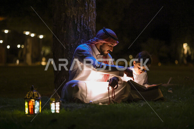 رجل عربي خليجي سعودي يعلم ابنه قراءة وتلاوة القرآن الكريم، تحفيظ القرآن للابناء، شهر رمضان المبارك، فانوس رمضان، إضاءة وزينة شهر رمضان، أجواء رمضانية، مناسبات دينية