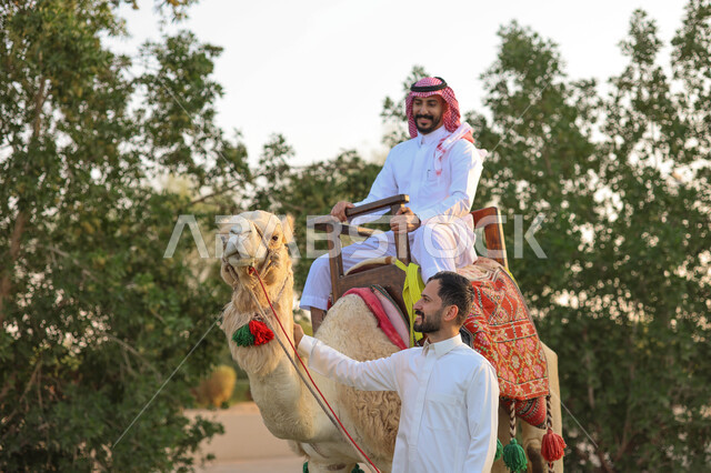 رجل عربي خليجي سعودي يمتطي الجمل العربي بجانبه رجل سعودي ، أنشطة ترفيهية ، فعالية سفاري موسم الرياض