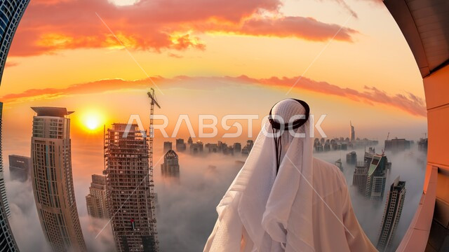 صوره من الخلف لرجل إماراتي عربي خليجي ينظر من الأعلى الى التطور العمراني في مدينة دبي أثناء الغروب، الأبراج وناطحات السحاب, المنشأت المعمارية, أجواء ضبابيه