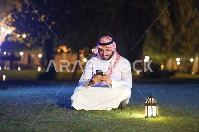 A Saudi Arabian Gulf man using a mobile phone, social networking, browsing websites, Ramadan lantern, the blessed month of Ramadan, lighting and decorations for the month of Ramadan, Ramadan atmosphere, religious occasions