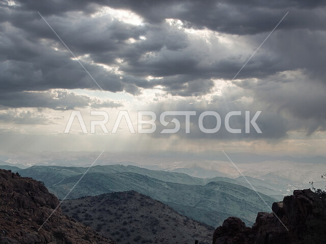 سماء غائمة تتخللها أشعة الشمس، جو ضبابي فوق سلسلة من المرتفعات الجبلية، اعشاب ونباتات جبلية