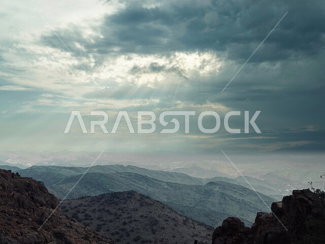 سماء غائمة تتخللها أشعة الشمس، جو ضبابي فوق سلسلة من المرتفعات الجبلية، اعشاب ونباتات جبلية