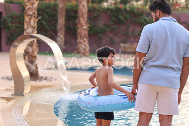 رجل عربي خليجي سعودي يقضي اوقات ممتعة مع ابنه، المشي على حدود البركة المائية، الاستعداد للسباحة في المسبح، اجواء صيفية ممتعة، رحلة سياحية ترفيهية، استخدام عوامة السباحة، عطلة صيفية سعيدة