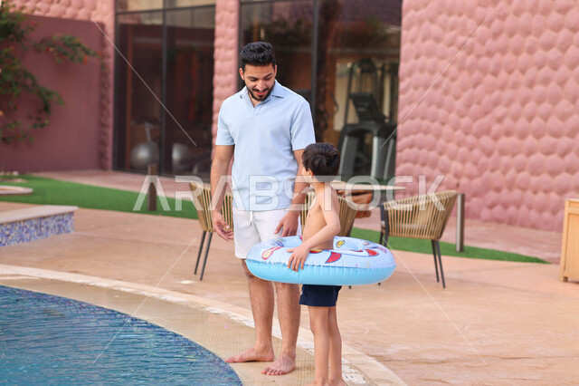 رجل عربي خليجي سعودي يقضي اوقات ممتعة مع ابنه، المشي على حدود البركة المائية، الاستعداد للسباحة في المسبح، اجواء صيفية ممتعة، رحلة سياحية ترفيهية، استخدام عوامة السباحة، عطلة صيفية سعيدة