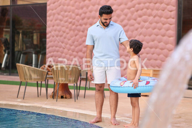 رجل عربي خليجي سعودي يقضي اوقات ممتعة مع ابنه، المشي على حدود البركة المائية، الاستعداد للسباحة في المسبح، اجواء صيفية ممتعة، رحلة سياحية ترفيهية، استخدام عوامة السباحة، عطلة صيفية سعيدة