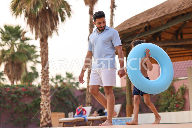 رجل عربي خليجي سعودي يقضي اوقات ممتعة مع ابنه، المشي على حدود البركة المائية، الاستعداد للسباحة في المسبح، اجواء صيفية ممتعة، رحلة سياحية ترفيهية، استخدام عوامة السباحة، عطلة صيفية سعيدة