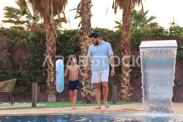 رجل عربي خليجي سعودي يقضي اوقات ممتعة مع ابنه، المشي على حدود البركة المائية، الاستعداد للسباحة في المسبح، اجواء صيفية ممتعة، رحلة سياحية ترفيهية، استخدام عوامة السباحة، عطلة صيفية سعيدة