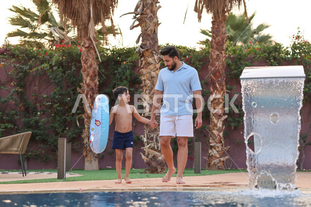 رجل عربي خليجي سعودي يقضي اوقات ممتعة مع ابنه، المشي على حدود البركة المائية، الاستعداد للسباحة في المسبح، اجواء صيفية ممتعة، رحلة سياحية ترفيهية، استخدام عوامة السباحة، عطلة صيفية سعيدة