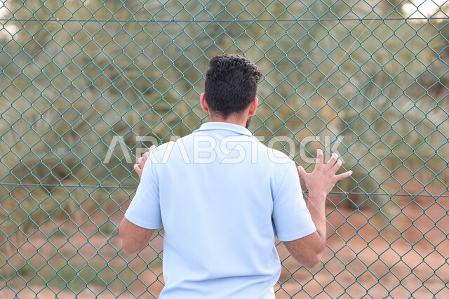 صورة من الخلف لرجل عربي خليجي سعودي يقف امام سياج حديد، شياج شبكي، اماكن عامة، الهواء الطلق، الاشجار والنباتات الخضراء