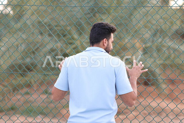 صورة من الخلف لرجل عربي خليجي سعودي يقف امام سياج حديد، شياج شبكي، اماكن عامة، الهواء الطلق، الاشجار والنباتات الخضراء