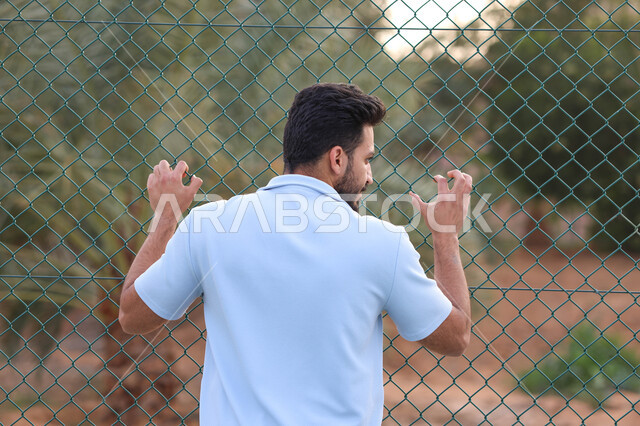 صورة من الخلف لرجل عربي خليجي سعودي يقف امام سياج حديد، شياج شبكي، اماكن عامة، الهواء الطلق، الاشجار والنباتات الخضراء