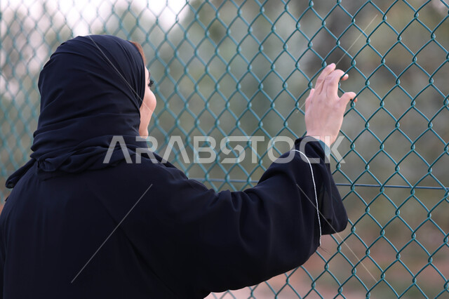 صورة من الخلف لامرأة عربية خليجية سعودية تقف امام سياج حديد، سياج شبكي، اماكن عامة، الهواء الطلق، الاشجار والنباتات الخضراء