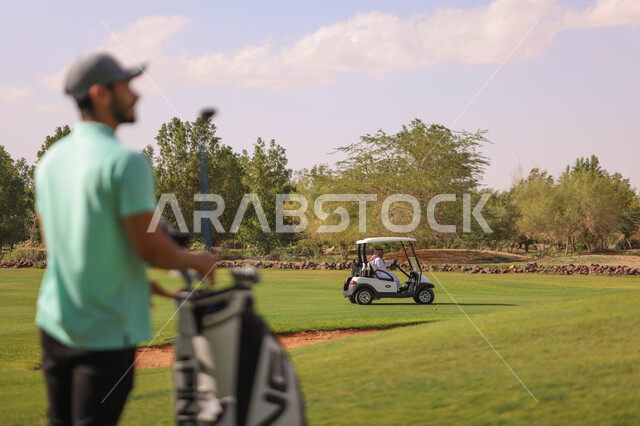 رجل عربي خليجي سعودي يحمل حقيبة الجولف، ممارسة لعبة الغولف على مساحات واسعة خضراء، أنشطة رياضية بالسعودية، لعبة الجولف، رياضات الألعاب الأولمبية الصيفية، أنشطة ترفيهية، سيارة كهربائية مختصة بنقل الركاب، ملعب الجولف