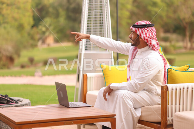رجل عربي خليجي سعودي يشير تحو شيء ما، استخدام الحاسوب المحمول، العمل عن بعد، اجهزة تقنية حديثة، تصفح الإنترنت، الجلوس في الهواء الطلق، الطبيعة الخضراء، جلسة عمل