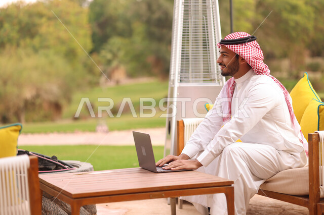 رجل عربي خليجي سعودي يستخدم الحاسوب المحمول، العمل عن بعد، إستخدام أجهزة تقنية حديثة، تصفح الإنترنت، الجلوس في الهواء الطلق، الطبيعة الخضراء، جلسة عمل