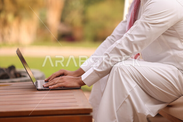 رجل عربي خليجي سعودي يستخدم الحاسوب المحمول، العمل عن بعد، إستخدام أجهزة تقنية حديثة، تصفح الإنترنت، الجلوس في الهواء الطلق، الطبيعة الخضراء، جلسة عمل