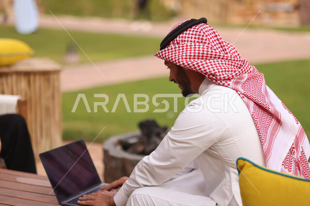 رجل عربي خليجي سعودي يستخدم الحاسوب المحمول، العمل عن بعد، إستخدام أجهزة تقنية حديثة، تصفح الإنترنت، الجلوس في الهواء الطلق، الطبيعة الخضراء، جلسة عمل
