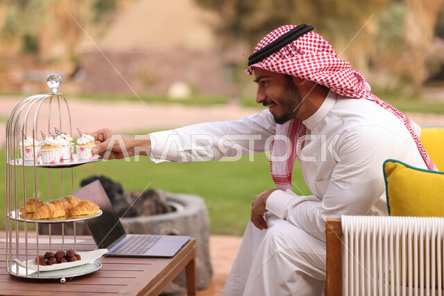 رجل عربي خليجي سعودي يستخدم الحاسوب المحمول، إنجاز مهام العمل، العمل عن بعد، تشيز كيك مع طبقات الكريمه، حلويات وسكريات، الاستمتاع باكل حلويات باردة، الجلوس في الهواء الطلق، الطبيعة الخضراء