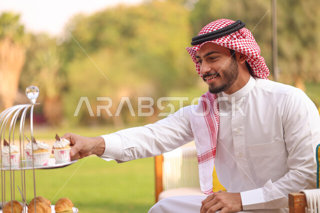 رجل عربي خليجيي سعودي يقضي أوقات ممتعة في الاماكن الترفيهية، تشيز كيك مع طبقات الكريمه، حلويات وسكريات، الاستمتاع باكل حلويات باردة، ايماءات وجه سعيدة، الجلوس في الهواء الطلق، الطبيعة الخضراء