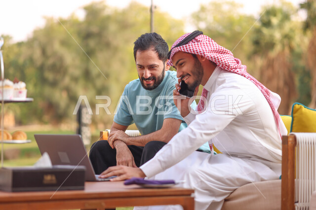 رجل عربي خليجي سعودي يجلس مع صديقه ويجري اتصال هاتفي، استخدام الحاسوب المحمول، العمل عن بعد، دردشة مع الأصدقاء، اجراء مكالمة هاتفية، الجلوس في الهواء الطلق، الطبيعة الخضراء