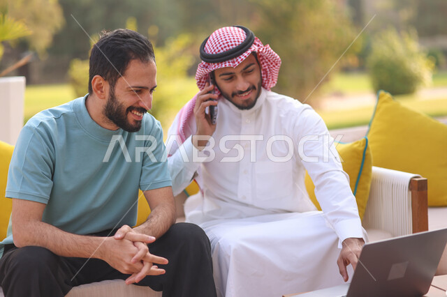 رجل عربي خليجي سعودي يجلس مع صديقه ويجري اتصال هاتفي، استخدام الحاسوب المحمول، العمل عن بعد، دردشة مع الأصدقاء، اجراء مكالمة هاتفية، الجلوس في الهواء الطلق، الطبيعة الخضراء